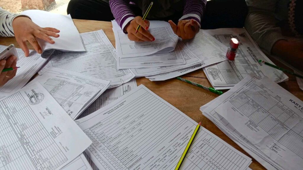 Indonesian payroll paperwork being reviewed manually, with multiple printed forms related to employee records, BPJS registration, and WLKP manpower reports spread across a wooden table.