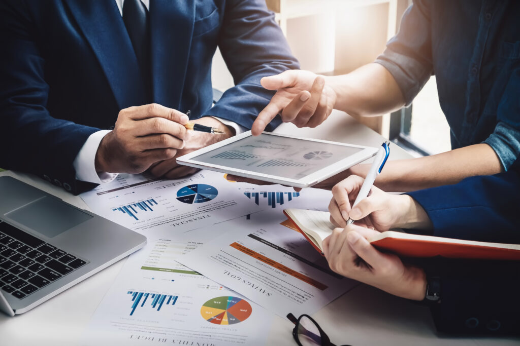 Three professionals review charts on a tablet and printed financial reports during an internal audit and compliance improvement meeting in an Indonesian service enterprise.